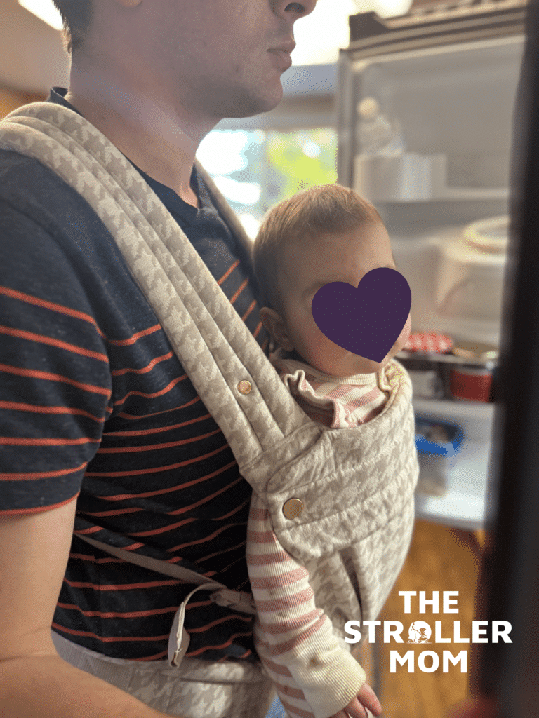 Dad wearing baby in the Mabe Monarch Carrier in houndstooth in the front inward-facing position at home, showing the carrier's adjustable fit for different body types