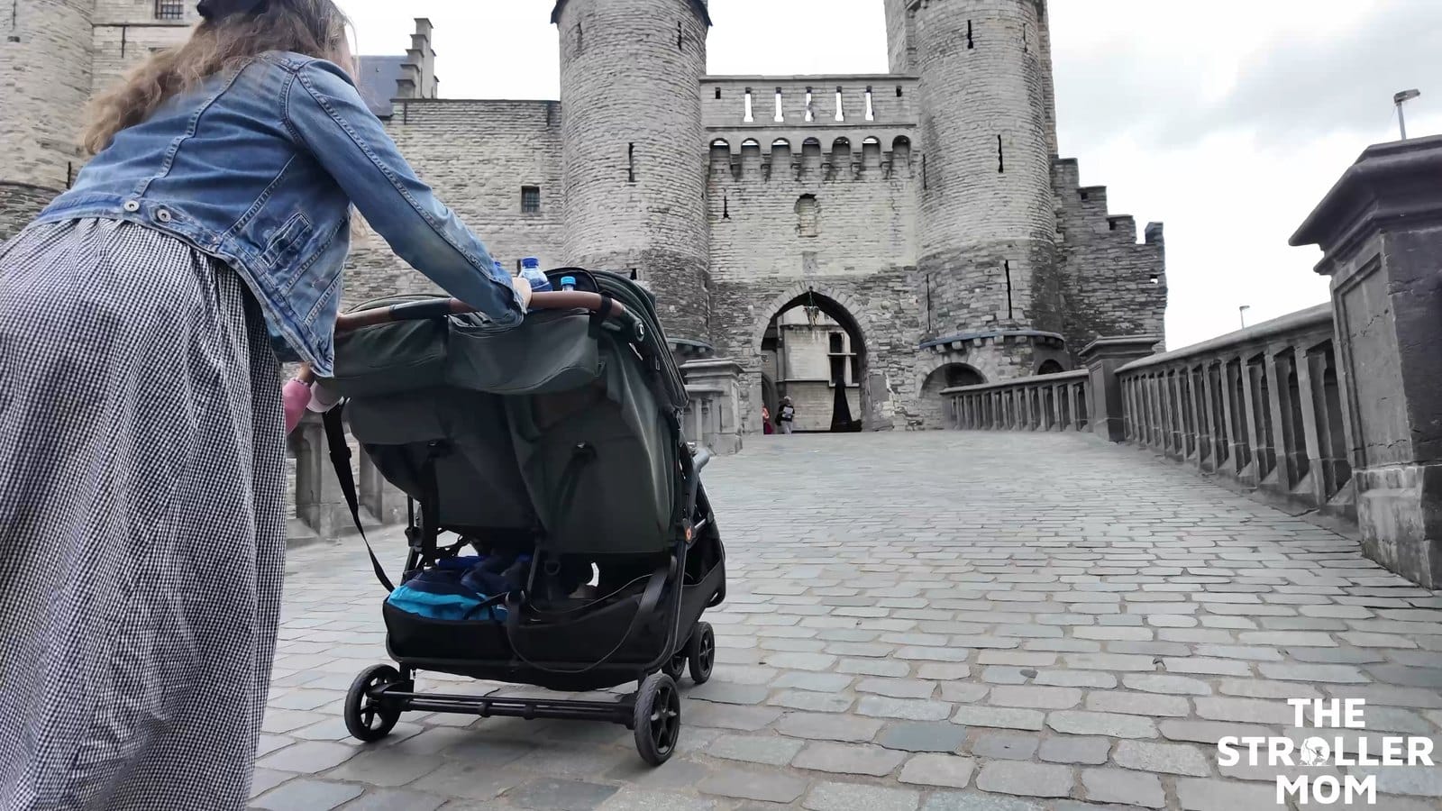 Rear view of Emily pushing the Zoe Twin V3 toward a stone castle, with outing essentials in the underseat basket.