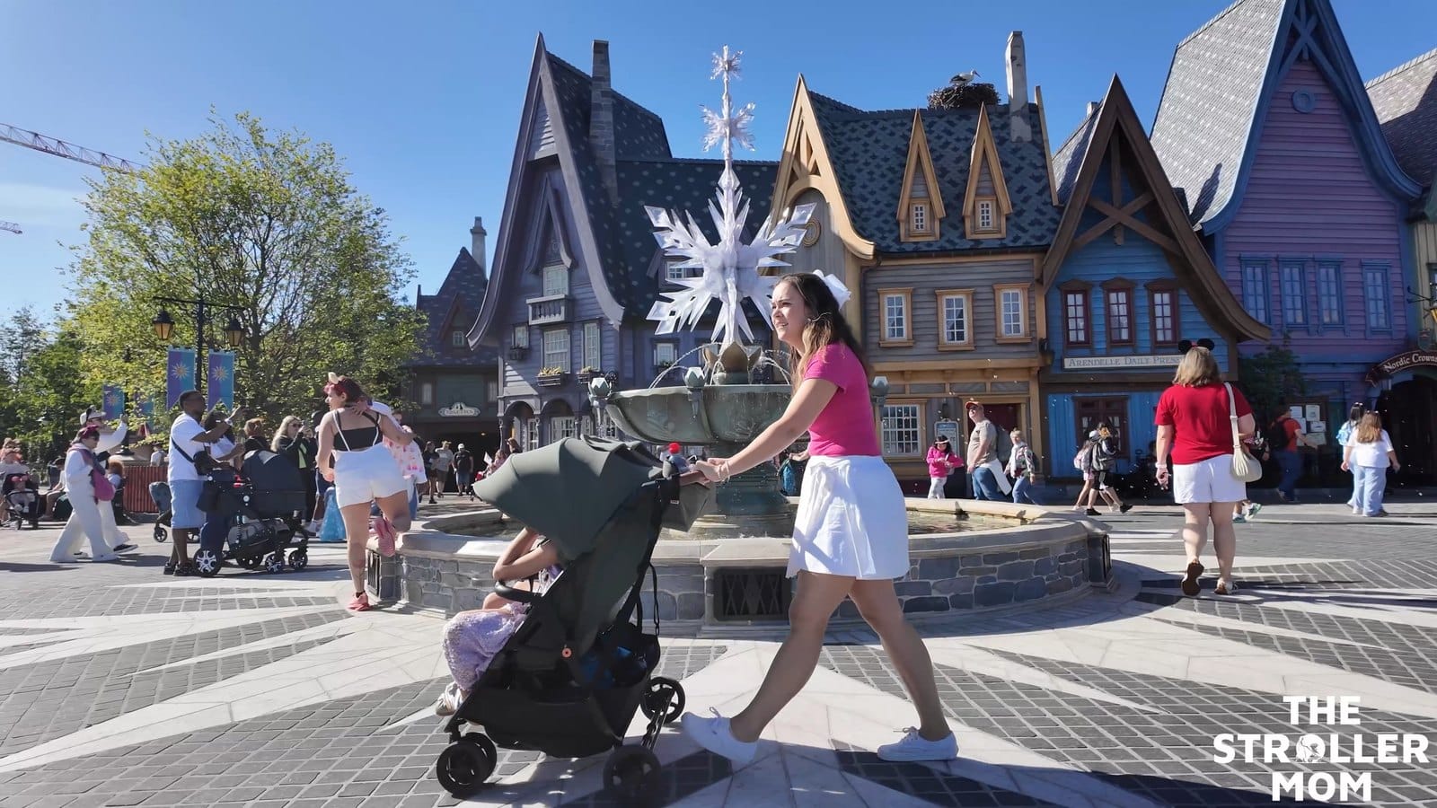 Emily pushing the Zoe Twin V3 past a fountain and colorful buildings during a theme park outing.